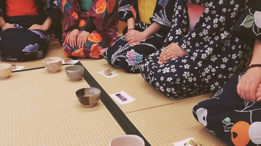 Students wearing yukata sit in a traditional Japanese room, holding tea bowls as they experience a Japanese tea ceremony.